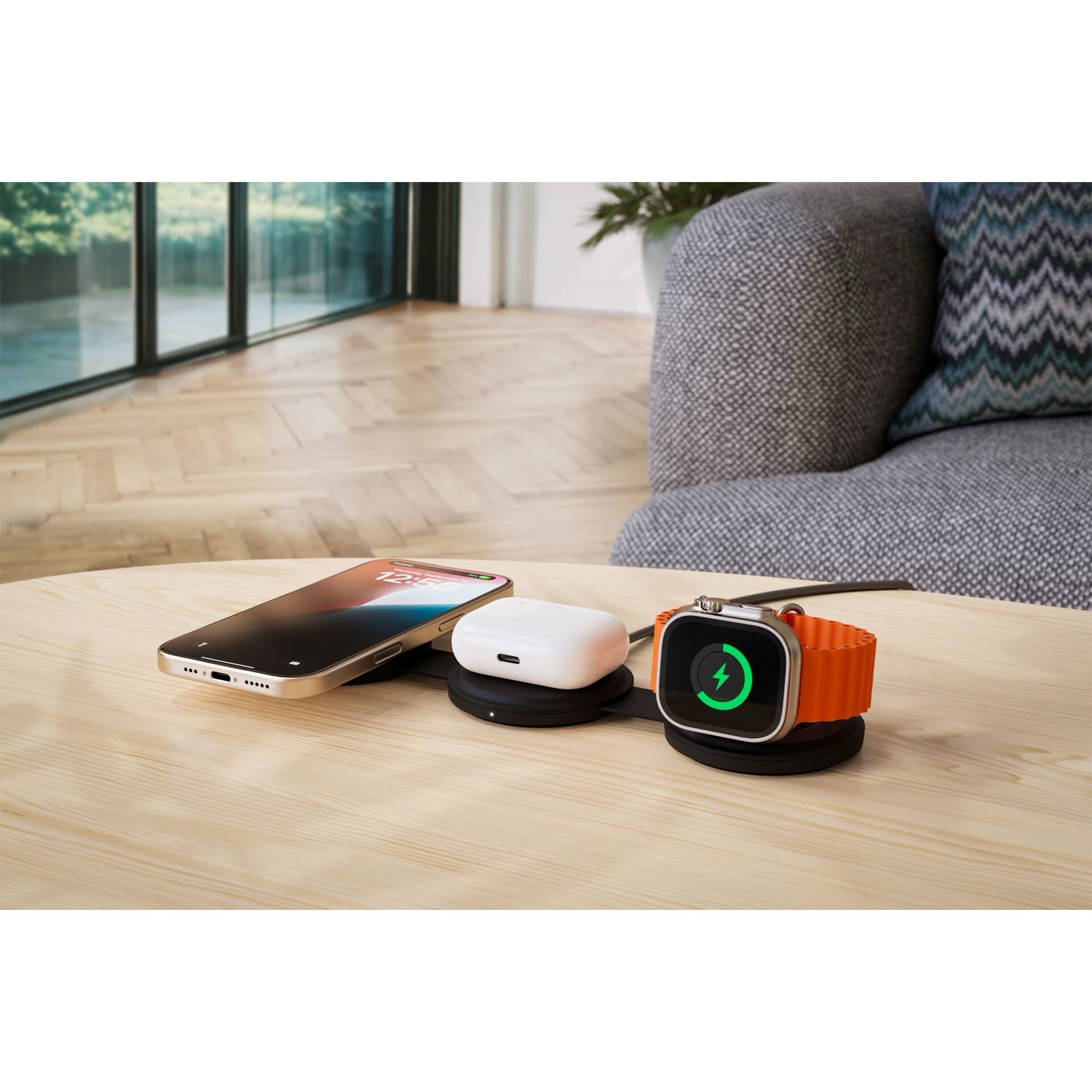 Ein Satechi OntheGo 3-in-1 Wireless Charger versorgt ein Smartphone, Ohrstöpsel und eine Smartwatch mit Qi2-Technologie auf einem Holztisch neben einer grauen Couch in einem modernen Wohnzimmer.