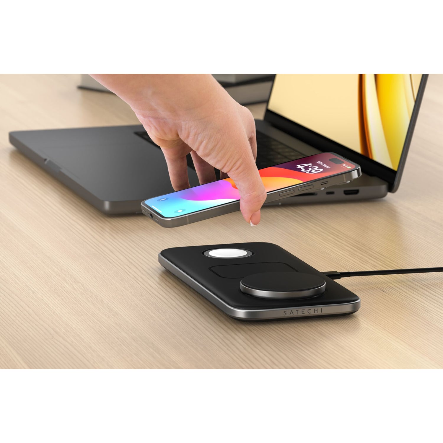 Eine Hand legt ein Smartphone auf das Satechi Qi2 Trio Wireless Charging Pad von Satechi, neben einem Laptop auf einem Holztisch.