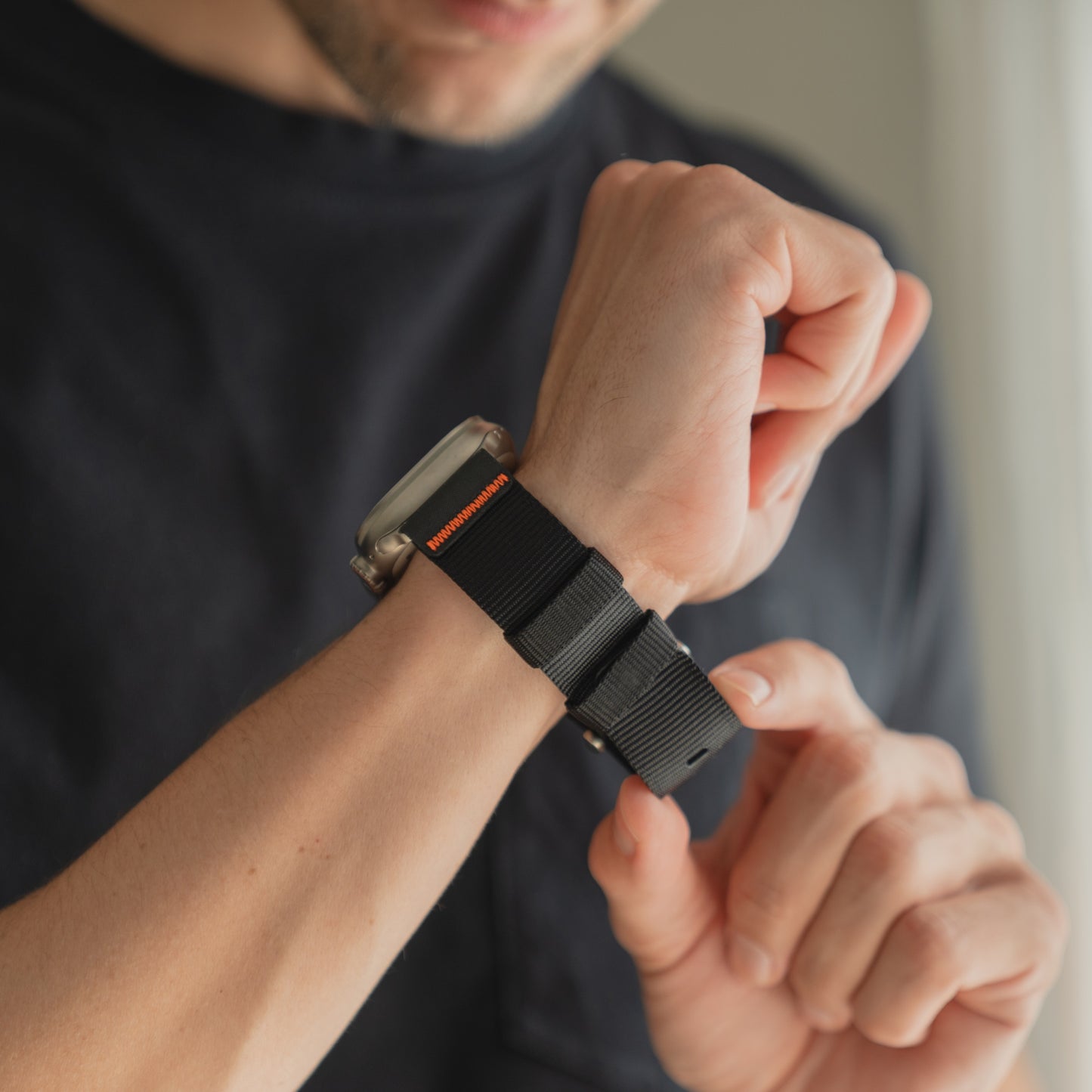 Jemand passt das arktisband Apple Watch Armband "Terra" von arktisband, ein 24 mm extrabreites Nylonband, an seine Apple Watch Ultra an, während er ein dunkles Hemd in einem Raum mit gedämpftem Licht trägt.