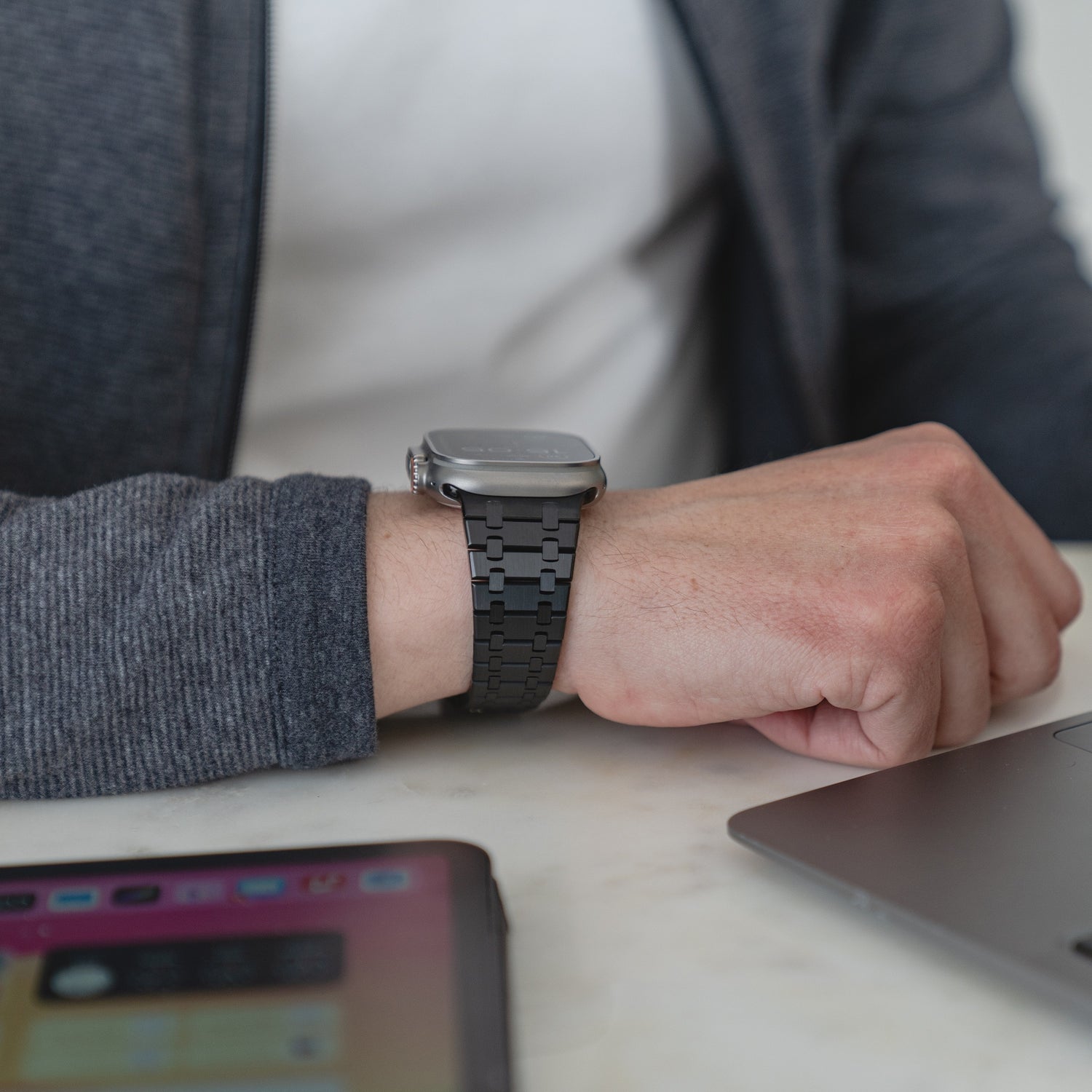 Eine Person, die ein Arktisband Edelstahlarmband „Symphony“ für die Apple Watch mit Butterfly-Schließe trägt, sitzt mit einem Laptop und einem Tablet an einem Tisch.
