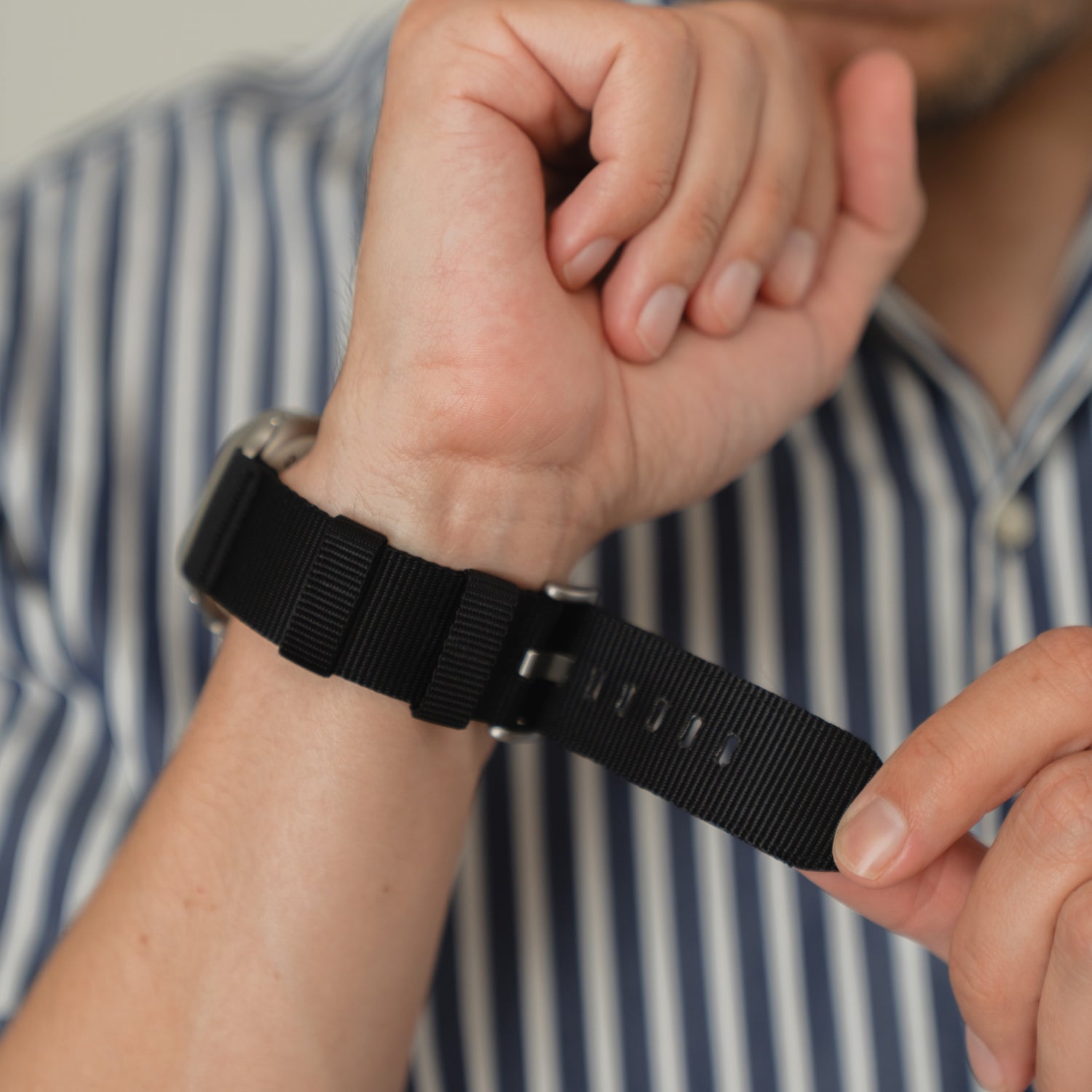 Eine Person passt das arktisband Apple Watch Armband "Terra" von arktisband mit einem extrabreiten 24-mm-Nylonband an ihr Handgelenk an, während sie ein blau-weiß gestreiftes Hemd trägt.