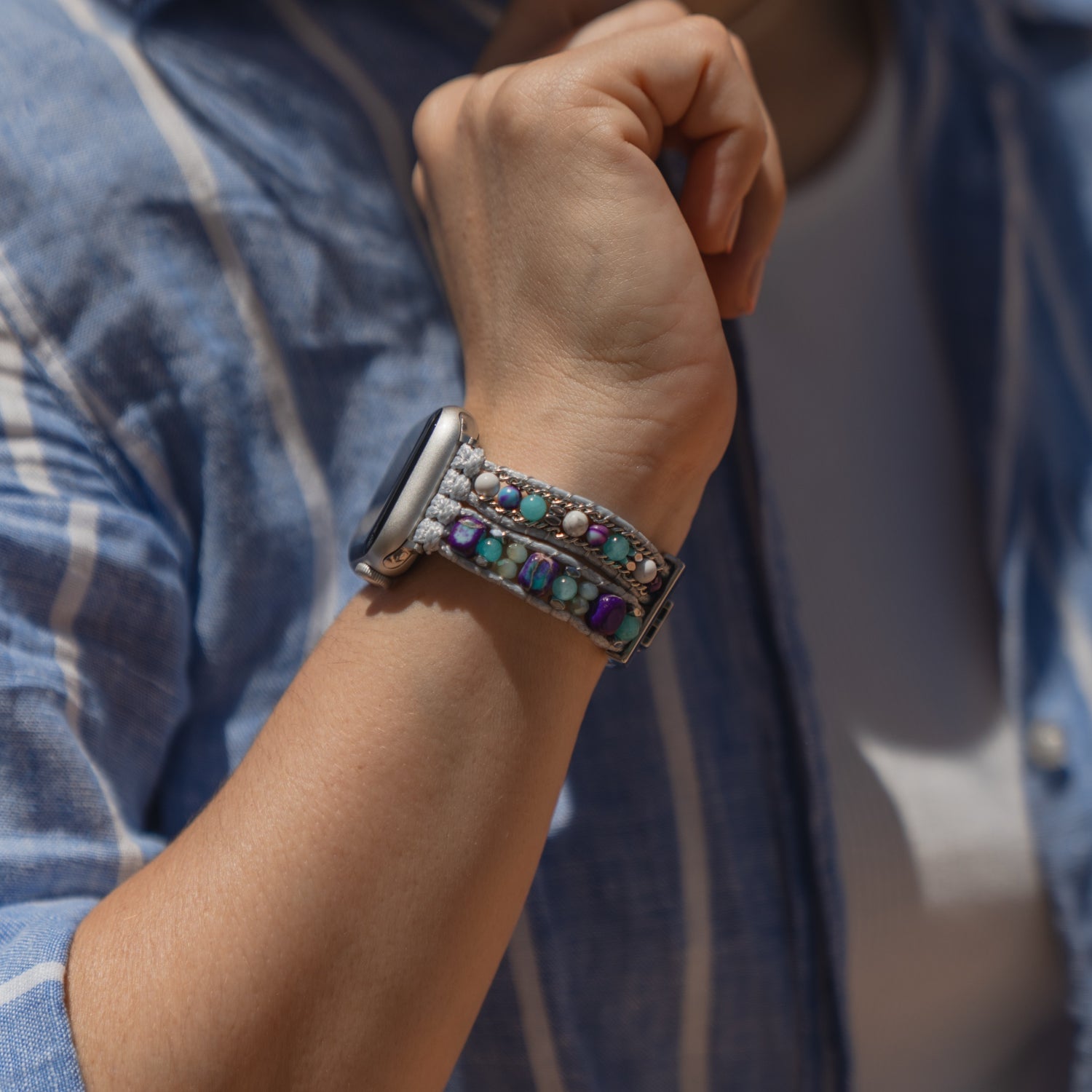 Eine Person, die ein „Sunset Spirit“-Armband von arktisband für die Apple Watch trägt – ein mehrfarbiges Perlenarmband –, zusammen mit ihrer Apple Watch und einem blau karierten Hemd.