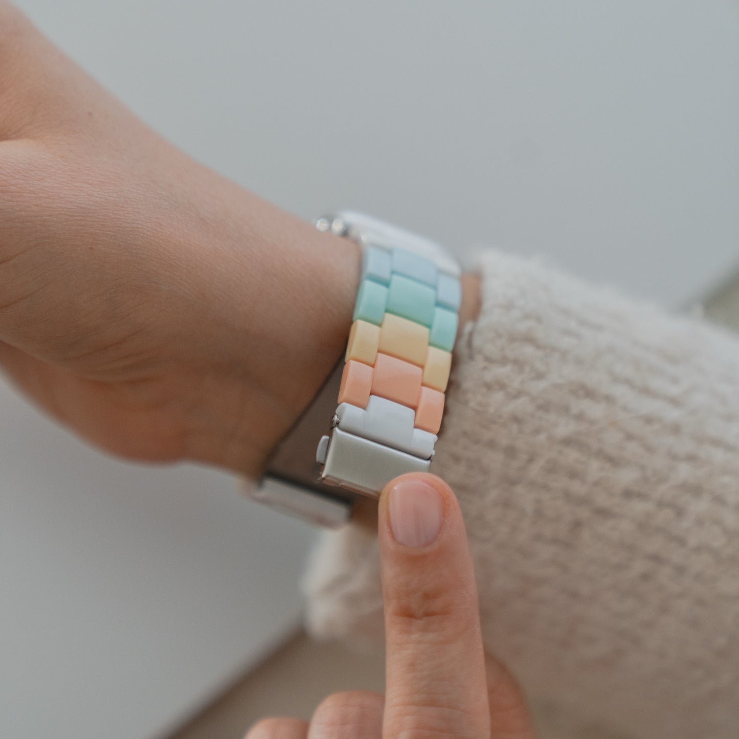 Eine Person zeigt auf das arktisband Apple Watch "Pixel Band", ein pastellfarbenes Armband im Retro-Look von arktisband, an ihrem Handgelenk, während sie einen cremefarbenen Pullover mit Struktur trägt.