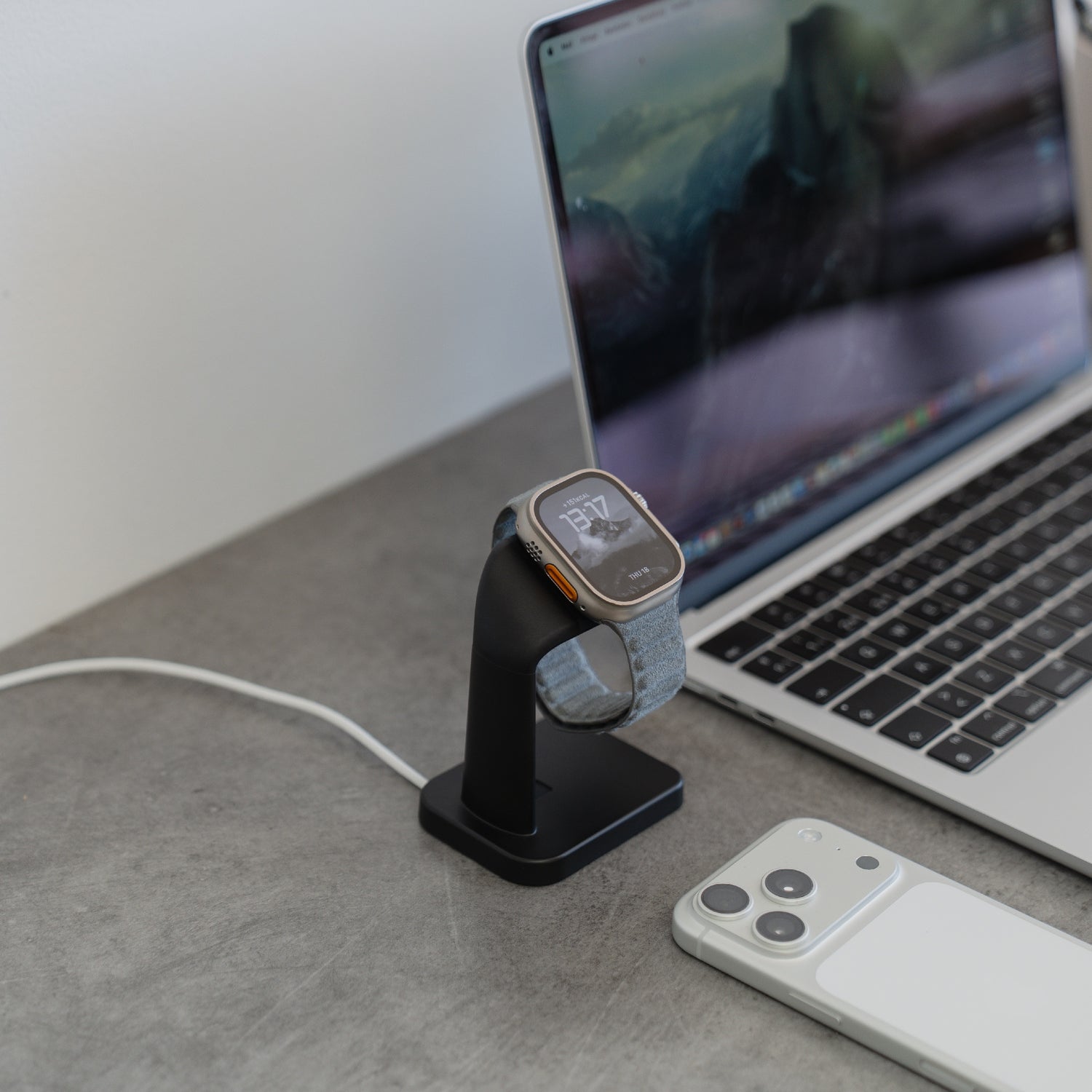 Ein ArktisPRO Apple Watch Charging Dock schwarz von ArktisPRO hält eine Smartwatch neben Laptop und Smartphone auf einem grauen Schreibtisch und schafft einen aufgeräumten, minimalistischen Design-Arbeitsplatz.