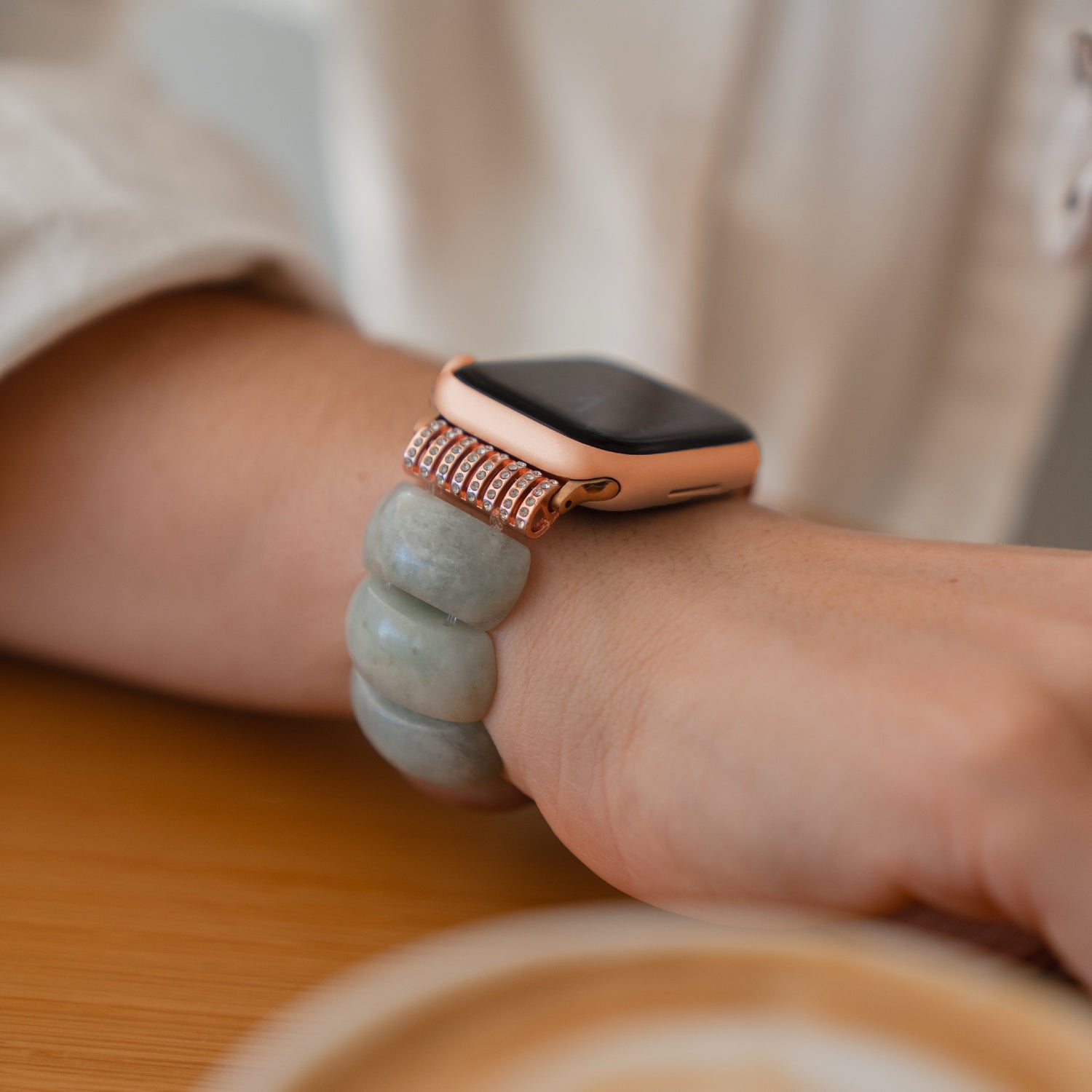 Die arktisband Apple Watch "Stone Pearl" von arktisband mit Naturstein-Armband und dehnbarem Gummizug-Verschluss wird am Handgelenk getragen, während sie auf einem Holztisch ruht.