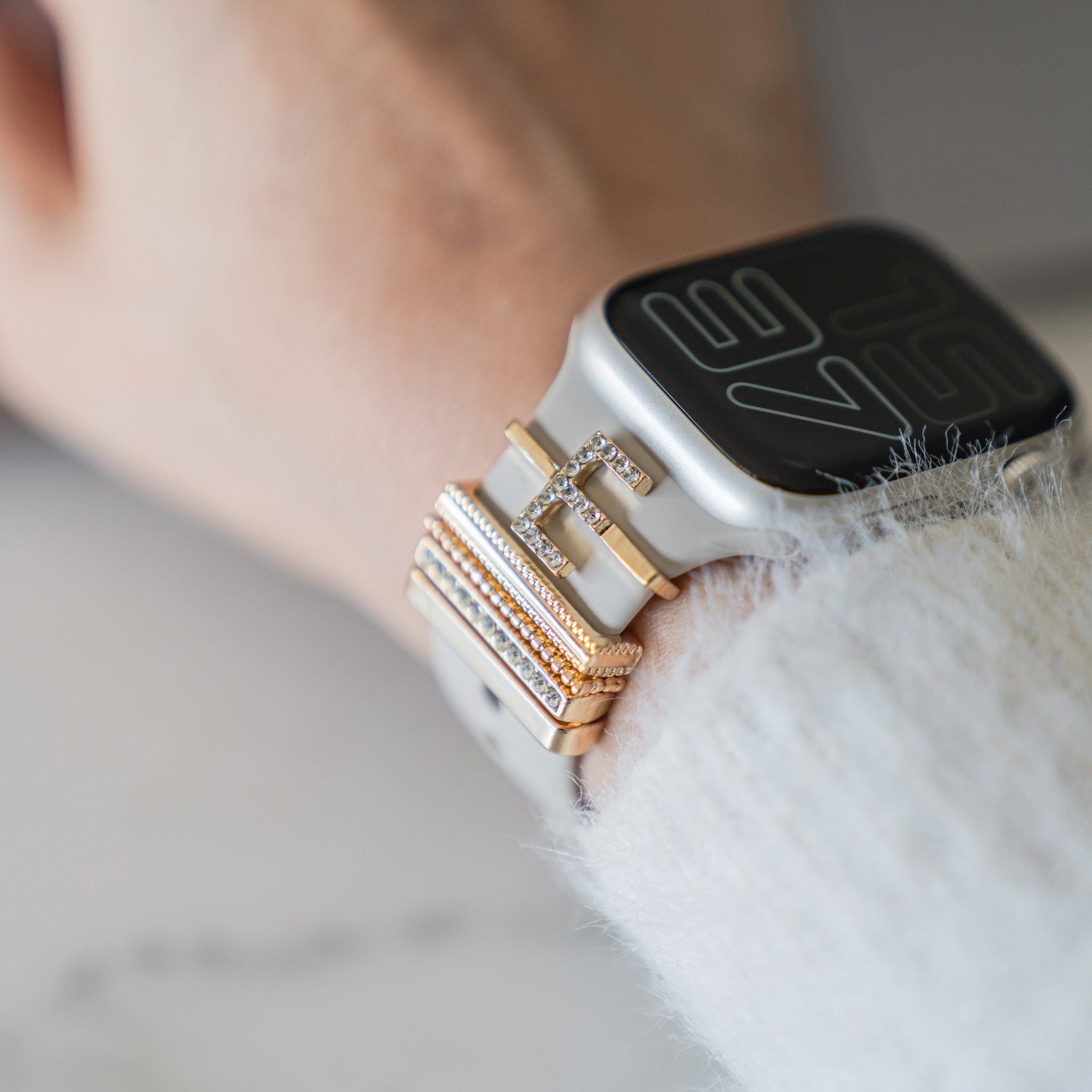 Nahaufnahme eines Handgelenks mit einer Smartwatch, die mit Gold- und Diamantarmbändern geschmückt ist, mit Arktisband Apple Watch Charms „E Style“ für ein elegantes Accessoire-Upgrade.