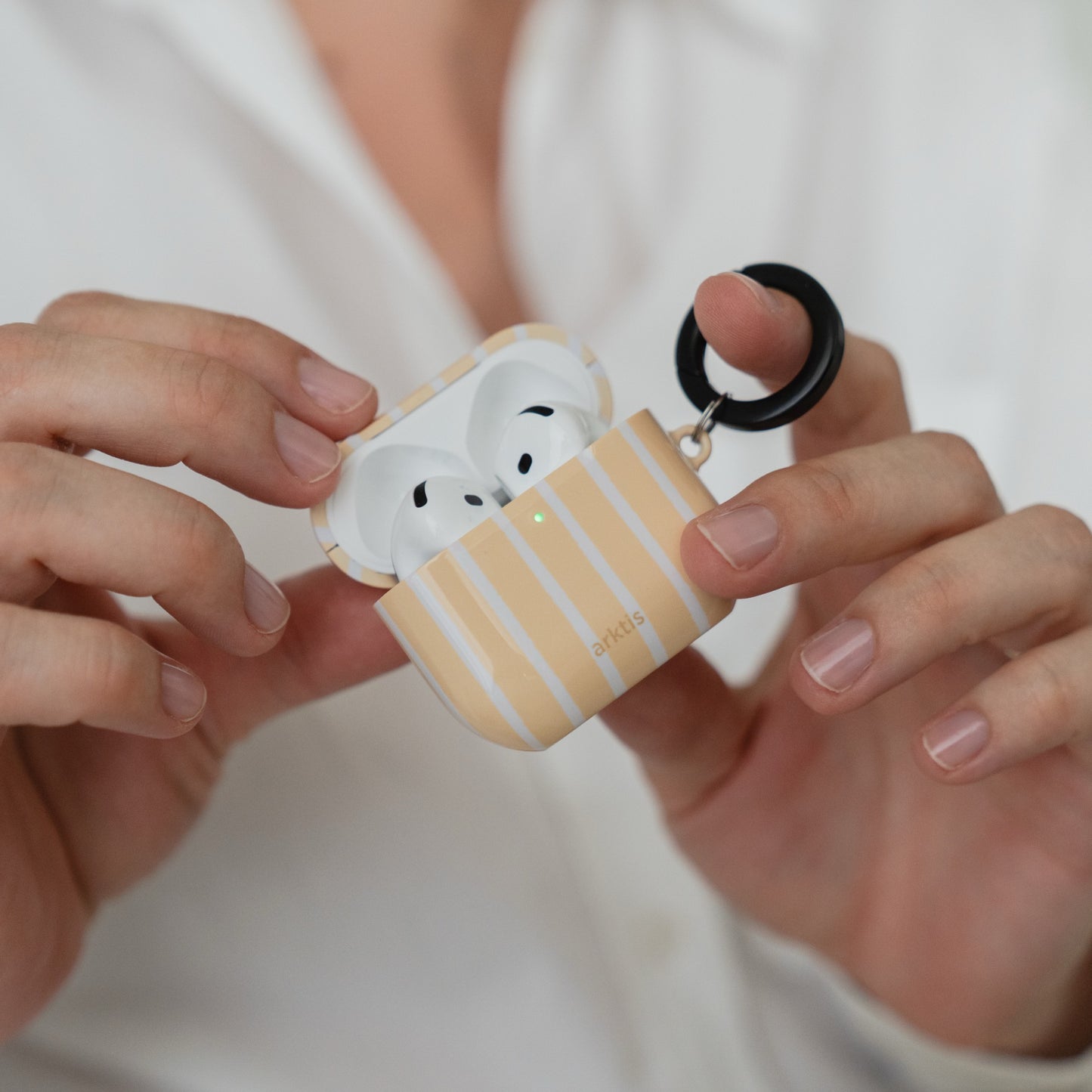 In den Händen wird eine geöffnete Arktis AirPods 5-Hülle – Sunny Stripes – gehalten, in der sich kabellose Ohrhörer mit einem schwarzen Befestigungsring befinden.