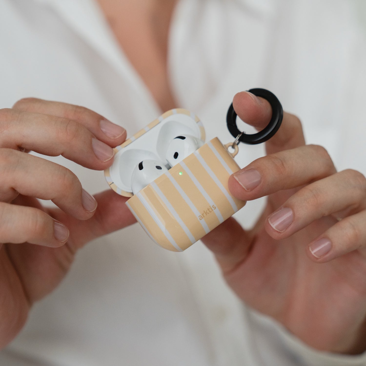 In den Händen wird eine geöffnete Arktis AirPods 5-Hülle – Sunny Stripes – gehalten, in der sich kabellose Ohrhörer mit einem schwarzen Befestigungsring befinden.