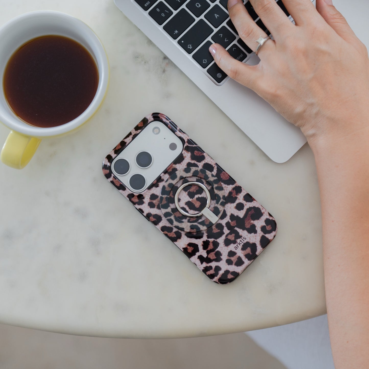 Eine Hand ruht auf einem Laptop, in einer gelben Tasse steht Kaffee, und ein Smartphone mit der „Arktis Magnetischer Ringhalter für MagSafe – Sweet Leopard“-Hülle – ausgestattet mit leistungsstarken N52-Magneten – rundet dieses stilvolle Ensemble an iPhone-Zubehör auf einem runden Tisch ab.
