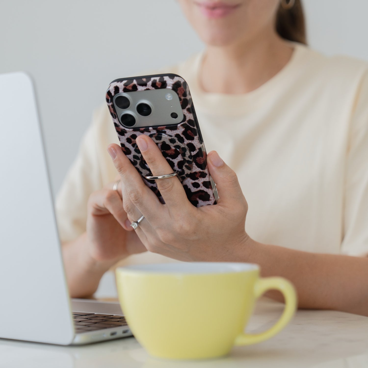 Eine Frau hält ihr Smartphone mit der „Arktis Magnetischer Ringhalter für MagSafe – Sweet Leopard“-Hülle in der Hand und sitzt an einem Tisch mit einem Laptop und einer gelben Tasse – perfekt für alle, die stilvolles iPhone-Zubehör lieben.