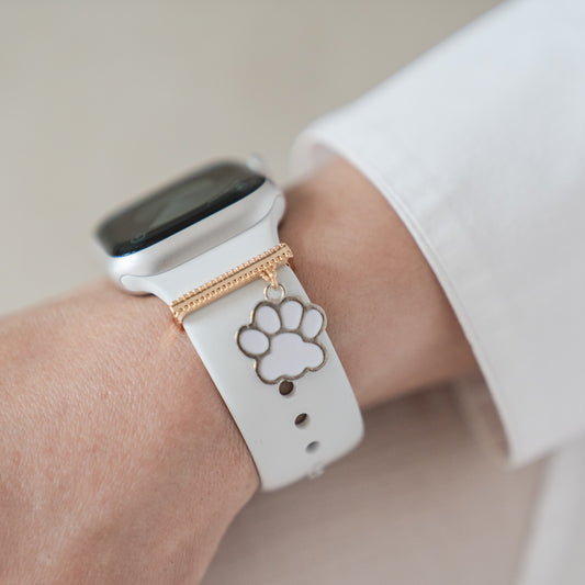 Ein Arktisband Apple Watch Charms „White Paw“ in Gold ziert ein weißes Arktisband Smartwatch-Armband, gezeigt an einem Handgelenk mit einer weißen Hülle.