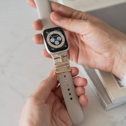 Eine Person hält eine Smartwatch mit Arktisband-Armband, verziert mit gestapelten Gold- und Diamant-Arktisband-Apple-Watch-Charms im „T-Stil“. Im Hintergrund liegt ein offenes Buch auf dem Tisch.