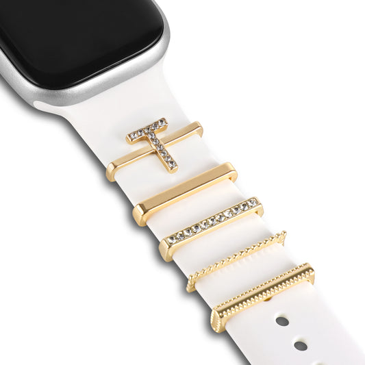 Eine Nahaufnahme einer Apple Watch mit einem weißen Arktisband, verziert mit fünf goldenen und strassbesetzten Arktisband-Apple-Watch-Charms im „T-Stil“ in Balkenform; oben links ist ein Teil des Zifferblatts sichtbar.