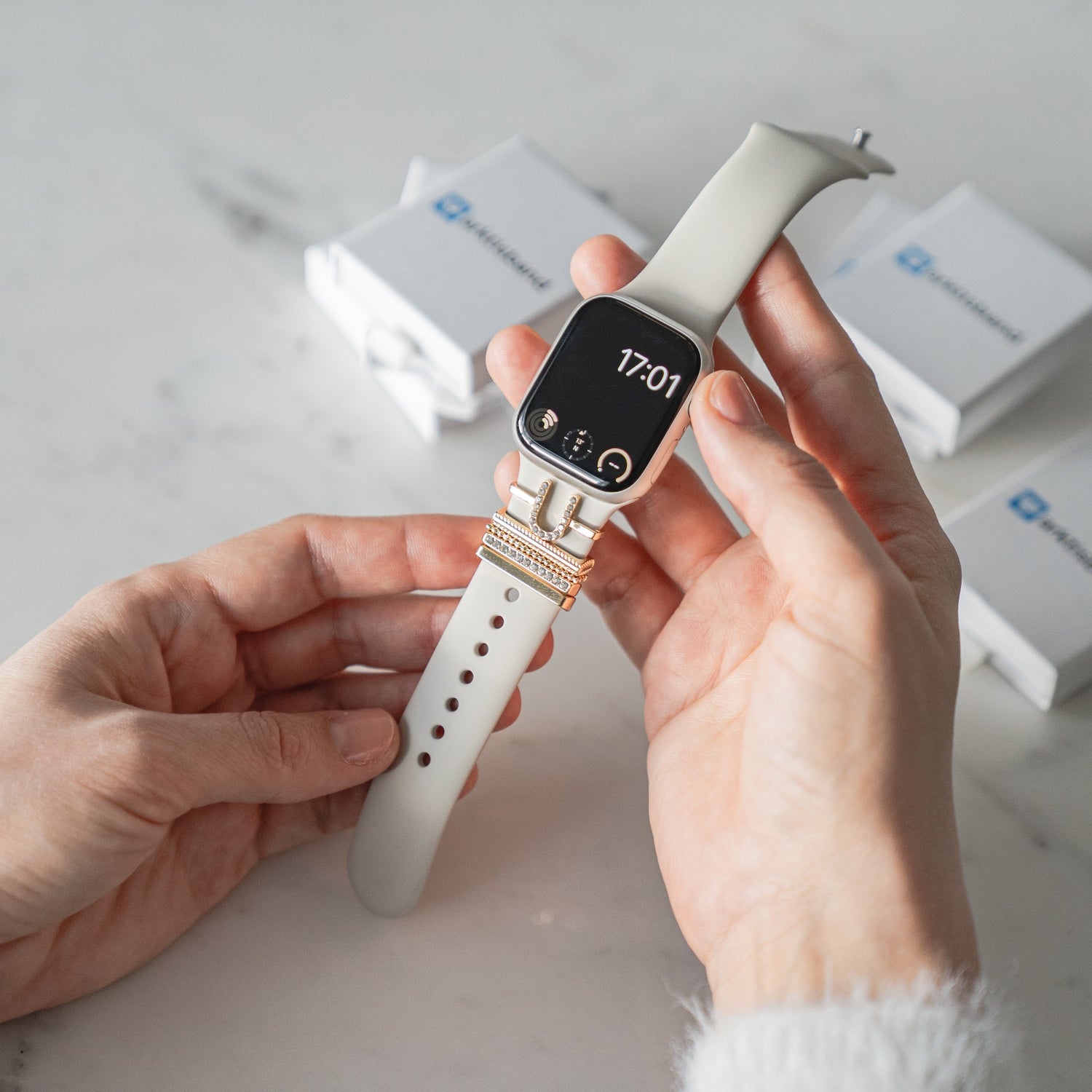 Eine Person hält eine arktisband Apple Watch Charms „U Style“ Smartwatch mit weißem Armband, goldenen Akzenten und der Uhrzeitanzeige 17:01 – ein Accessoire von arktisband für jeden Anlass.