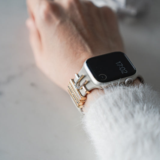 Ein mit Arktisband Apple Watch Charms „U Style“ und goldenen Armbändern gestyltes Handgelenk, gepaart mit einem flauschigen weißen Pullover, glänzt auf einer hellen Oberfläche.