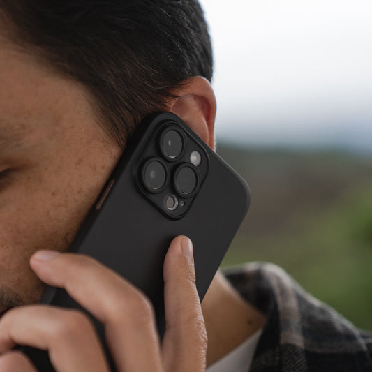 Eine Person hält ein iPhone 15 in einer schwarzen ArktisPRO iPhone 15 Hülle ULTRADÜNN von ArktisPRO mit Dreifachkamera ans Ohr, als würde sie telefonieren. Ihr Gesicht ist teilweise vor unscharfem Hintergrund sichtbar.