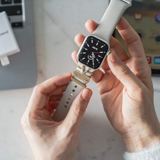 Jemand befestigt eine weiße Smartwatch mit einem goldenen Verschluss und fügt arktisband Apple Watch Charms „V Style“ von arktisband für einen extravaganten Look hinzu, neben einem Laptop und einem Tablet auf dem Schreibtisch.