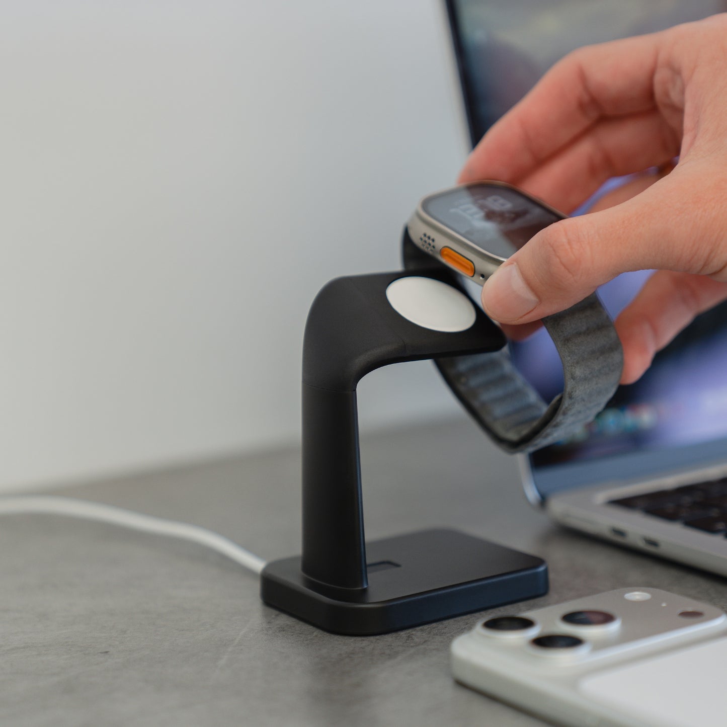 Eine Hand platziert eine Apple Watch auf das ArktisPRO Apple Watch Charging Dock schwarz von ArktisPRO mit cleverer Kabelführung neben Smartphone und Laptop - alles im minimalistischen Design.