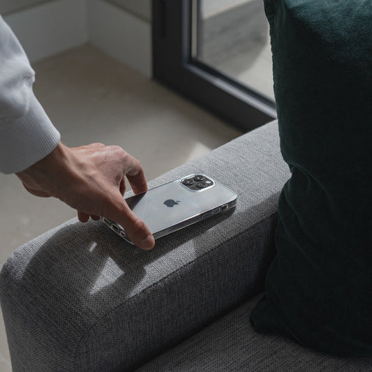 Eine Hand nimmt ein Arktiscase Arktis iPhone 15 Pro ZERO Case Ultra Clear von einem grauen Sofa neben einem grünen Kissen und einem Fenster.