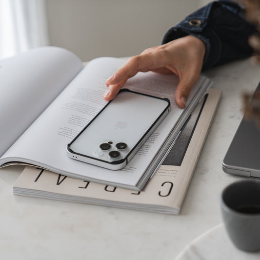 Eine Hand ruht auf einem offenen Buch, auf dem sich der Aviato AirZERO Alu Bumper für das iPhone 14 Pro von Aviato befindet, bereit zum kabellosen Laden. Daneben stehen ein geschlossenes Buch und eine graue Kaffeetasse auf einem weißen Tisch.