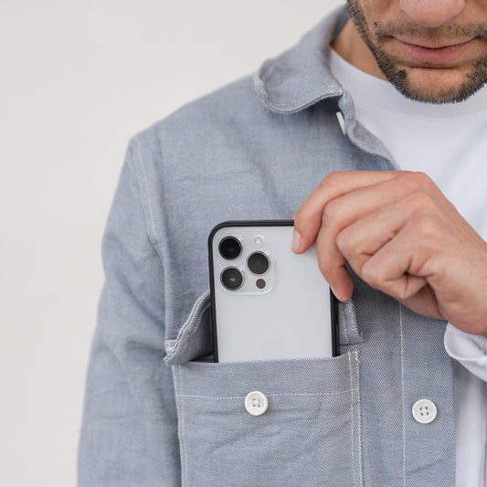 Ein Mann steckt ein iPhone 14 Pro Max mit drei Kameras, geschützt durch den Aviato AirZERO Alu Bumper von Aviato, in die Brusttasche seines hellblauen Hemdes.
